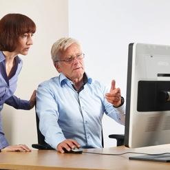 Frau und Mann betrachten gemeinsam einen Computerbildschirm an einem Holztisch. Der Mann erklärt etwas.