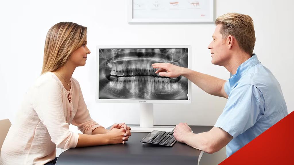 Zahnarzt zeigt Patientin auf Röntgenbild am Monitor, beide sitzen an einem Tisch in einer modernen Praxis.